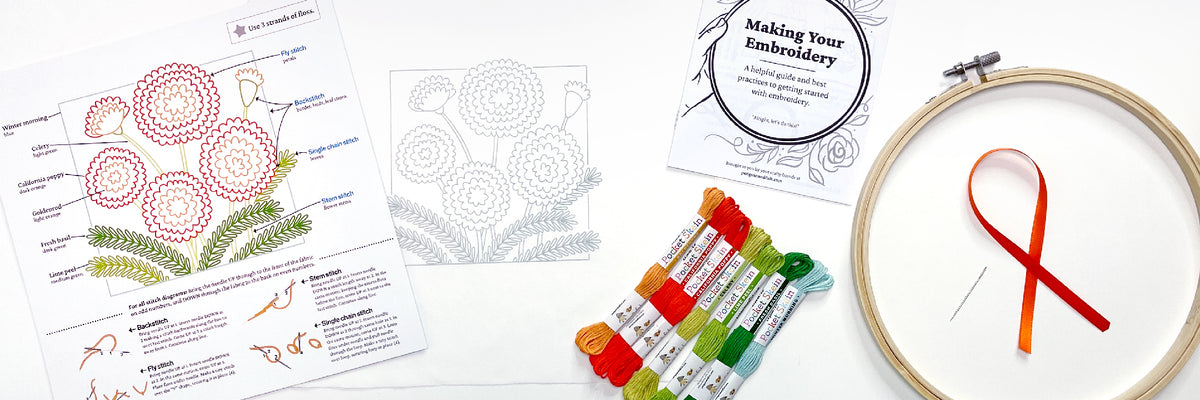 a white surface with the contents of the Marigold embroidery kit from Penguin & Fish. Includes the instructions, fabric with design printed on it, embroidery floss, embroidery instruction booklet, hoop, ribbon, and needle